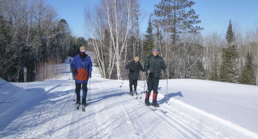 Three people on cross country skis slide over a snowy road. 